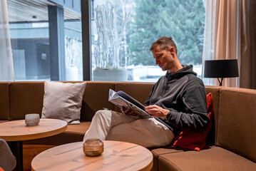 Wall Mural - Young man reading travel catalog while relaxing in lounge. Male is looking through magazine while sitting on sofa. He is resting in lobby at luxurious resort.