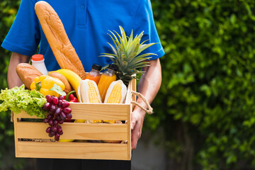 Asian man farmer wears delivery uniform he holding full fresh vegetables and fruits in crate wood box in hands ready give to customer harvest organic food on the garden place green leaves background