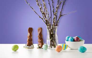 Wall Mural - easter, holidays and object concept - pussy willow branches, colored eggs and chocolate bunnies on table over violet background