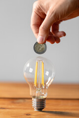 Wall Mural - electricity, energy crisis and power consumption concept - close up of hand with coin and lightbulb on wooden table