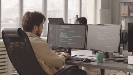 Rear view slowmo of male programmer typing sitting in front of two computer monitors in office typing program code on keyboard while developing computer game