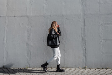 Wall Mural - Fashionable beautiful girl model in young stylish clothes with a leather jacket, jeans and boots with a bag puts on sunglasses and walks near a gray wall on a sunny day