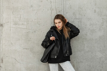 Canvas Print - Fashion young beauty woman hipster model with rock leather jacket, hoodie and fashion leather bag stands near a concrete gray wall. Female urban style, fashion and beauty