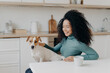 Playful woman with Afro haircut, pets her breed dog, have fun together, pose in cozy kitchen, drink coffee, laugh happily