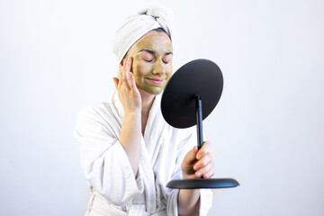 Canvas Print - Young woman with a green mask on her face in a bathrobe in front of a mirror.