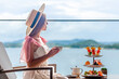Young lady in white dress drinking tea on Afternoon tea with mini canape and selection of various sweets with sea on background. Elegant woman enjoy ocean view, eating desserts in luxury restaurant.