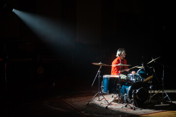 Young man rehearsing on drum set in spotlight in dark studio. Caucasian rock musician recording drums in hall. Drummer training concept