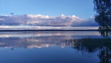 The Branches Of A Birch Tree Overhang The Water. Reeds Grow In The Water Near The Shore Of The Lake And There Are Ripples. On The Opposite Shore Is A Forest. The Blue Sky With Clouds Is Reflected 