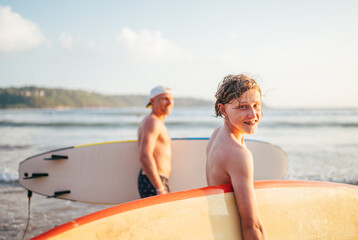 Wall Mural - Smiling teen boy with surfboard go to the sea for surfing. He have a winter vacation and enjoying a beautiful sunset light with father on Sri Lanka island. Family active vacation concept.