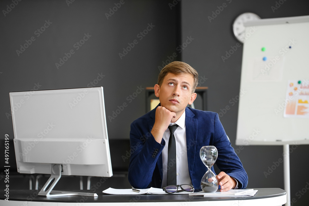 Thoughtful young businessman with hourglass sitting at table in office
