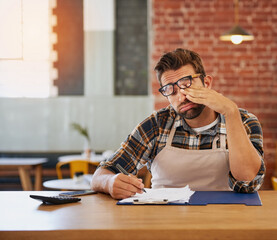 This admin will be the end of me. Shot of an exhausted young business owner doing admin at a table in his coffee shop.