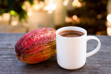 Wall Mural - Cup of hot chocolate drink and fresh cocoa fruit on wooden table with blurred background.