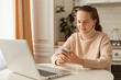© sementsova321 - Image of delighted smiling adorable woman wearing beige jumper posing in kitchen and using cell phone, holding smart phone in hands and typing, answering messages.
