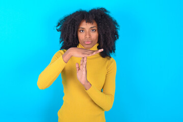 Wall Mural - Young woman with afro hairstyle wearing yellow turtleneck over blue background being upset showing a timeout gesture, needs stop, asks time for rest after hard work, demonstrates break hand sign