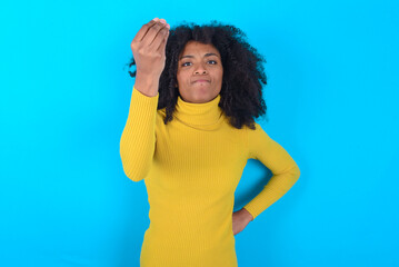 Wall Mural - Young woman with afro hairstyle wearing yellow turtleneck over blue background angry gesturing typical italian gesture with hand, looking to camera