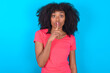 © Roquillo - Surprised Young girl with afro hairstyle wearing pink t-shirt over blue background makes silence gesture, keeps finger over lips and looks mysterious at camera