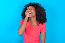 Young Girl With Afro Hairstyle Wearing Pink T-shirt Over Blue Background Makes Face Palm And Smiles Broadly, Giggles Positively Hears Funny Joke Poses