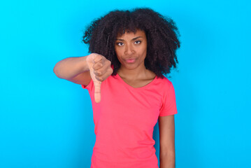 Wall Mural - Young girl with afro hairstyle wearing pink t-shirt over blue background looking unhappy and angry showing rejection and negative with thumbs down gesture. Bad expression.
