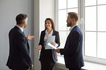 Business professionals. Smiling business people discuss work, project strategy and exchange ideas together. Two men and woman in business clothes stand with documents in hand near window in office.