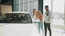 African American Man And Caucasian Woman While Walking At Modern Auto Center. Happy Couple Choosing New Vehicle Together. Beautiful Couple Standing At The Car Dealership And Making Their Decision.