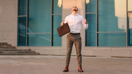 Successful lucky happy arabic spanish business man boss leader candidate entrepreneur with briefcase standing outdoors near modern company building wins gets opportunity new job makes victory gesture