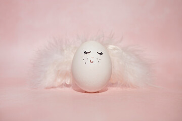 Wall Mural - An egg with a funny face and angel wings on a pink background. Easter concept.