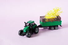 A Green Toy Tractor Is Carrying Yellow Seedlings In A Trailer. Spring Planting And Agricultural Work. Pastel Pink Background. Copy Space