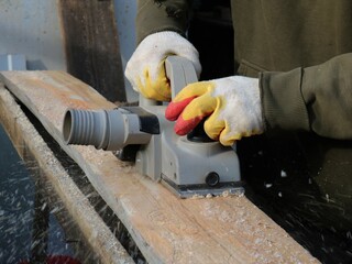 a carpenter with an electric planer in the process of working on wood, scraping off the top layer of a wooden board with a planer in the hands of a carpenter with sawdust flying to the side