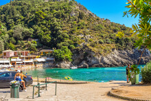 A Couple Riding A Motorcycle Park Near A Small Cove On Paleokastritsa Beach, With Shops And Cafes In View On A Summer Day On The Island Of Corfu, Greece. 