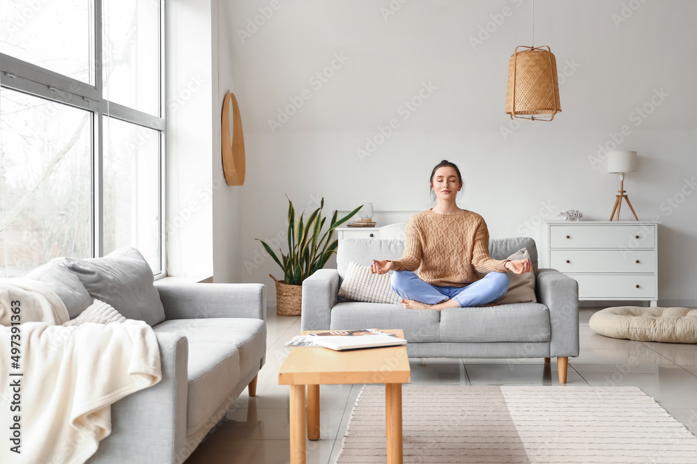 Beautiful young woman meditating on couch at home