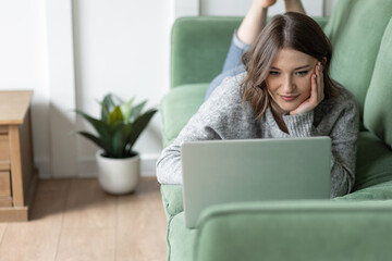 Wall Mural - Young beautiful smiling woman using smart phone lying on the couch. Concept of freelance job, home office. Female using technology for learning, shopping, e-commerce, communication, social media