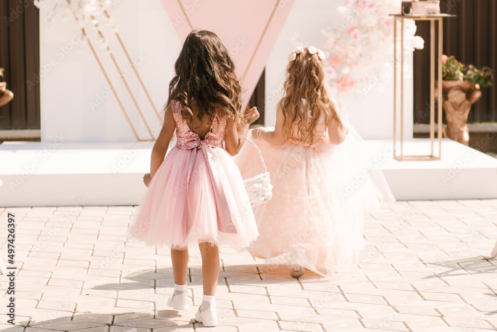 Little beautiful girls in puffy dresses eat petals and rings at the wedding, back view