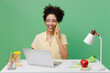 © ViDi Studio - Young smiling cheerful employee business woman of African American ethnicity in shirt sit work at white office desk with pc laptop talk speak on mobile cell phone isolated on plain green background