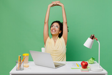 Wall Mural - Young employee business woman of African American ethnicity wear shirt sit work at white office desk with pc laptop stretch hands rest isolated on plain green background. Achievement career concept