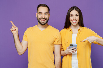 Wall Mural - Young happy couple two friends family man woman together in yellow casual clothes hold use mobile cell phone point index finger aside on workspace isolated on plain violet background studio portrait.