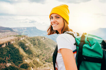 Wall Mural - Back view young happy smiling woman backpacker with brunette hair relaxation on top rocky mountains, with backpack, yellow hat. Leisure after walking valley. Adventure, travel, holiday concept.