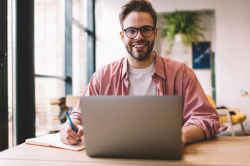 Wall Mural - Cheerful male freelancer with education textbook smiling at camera during laptop programming, portrait of happy IT professional with digital netbook enjoying remote working in cafe interior