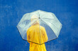 Mid waist portrait of unrecognizable woman in yellow waterproof clothes under rain with umbrella. Horizontal rear view of person outdoors holding umbrella on blue background. Weather concept