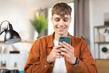 Close Up Of Smiling Young Man Sitting At Table And Using Modern Smartphone. Young Man In Brown Shirt Sitting At Modern Home And Serfing Social Networks Enjoying Leisure Time.