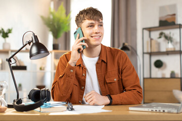 Wall Mural - Pleasant smiling Caucasian businessman talking on mobile and working on laptop at table. Young man in casual wear sitting at workplace and using modern gadgets.