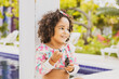 © EZ PHOTOS - Portrait of smiling baby girl with afro hair style and sunglasses in a colourful swimsuit outfit blowing a bubble outdoors. Afro-American and Afro-Brazilian children summer concept with copy space.
