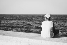 Closeup Shot Of A Boy Sitting On The Stone And Looking At The Waves Of A Sea In Black And White