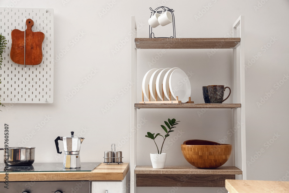 Modern shelving unit with tableware and coffee maker on stove near white wall in kitchen