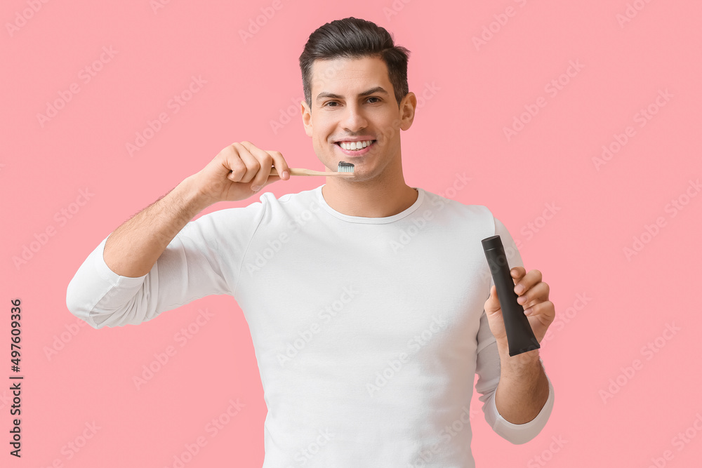 Handsome young man brushing teeth with activated charcoal tooth paste on pink background
