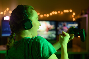 Wall Mural - Games online, a young girl playing computer games using a joystick or a gamepad. On three large monitors, creative light. Strategies, racing and gaming business. Creative green light.