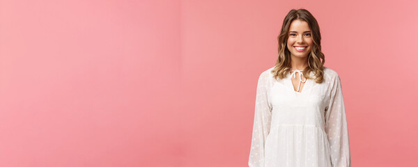 Spring, beauty and women concept. Portrait of tender feminine blonde girl with white beaming smile standing in light cute dress over pink background with happy attitude, positive emotions