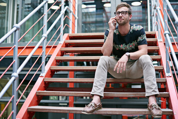 Sticker - Staying in touch with his clients. Shot of a handsome young man using his mobile phone while sitting on the steps of an office.