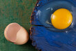 A piece of cracked wood painted blue. On it lies a raw, broken egg, with egg shells next to it. Shades of green and orange in the background, abstract background. Close-up.