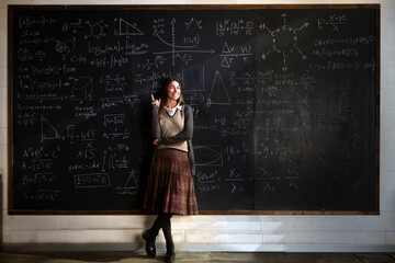 Cinematic shot of young female teacher or student indicates with forefinger at blackboard with math formulas and equations and smiles in camera in college or university classroom.
