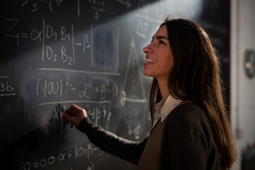 Cinematic shot of young female teacher is writing math formulas and equations with chalk on blackboard to explain educational material task to her pupils or students in college or university classroom